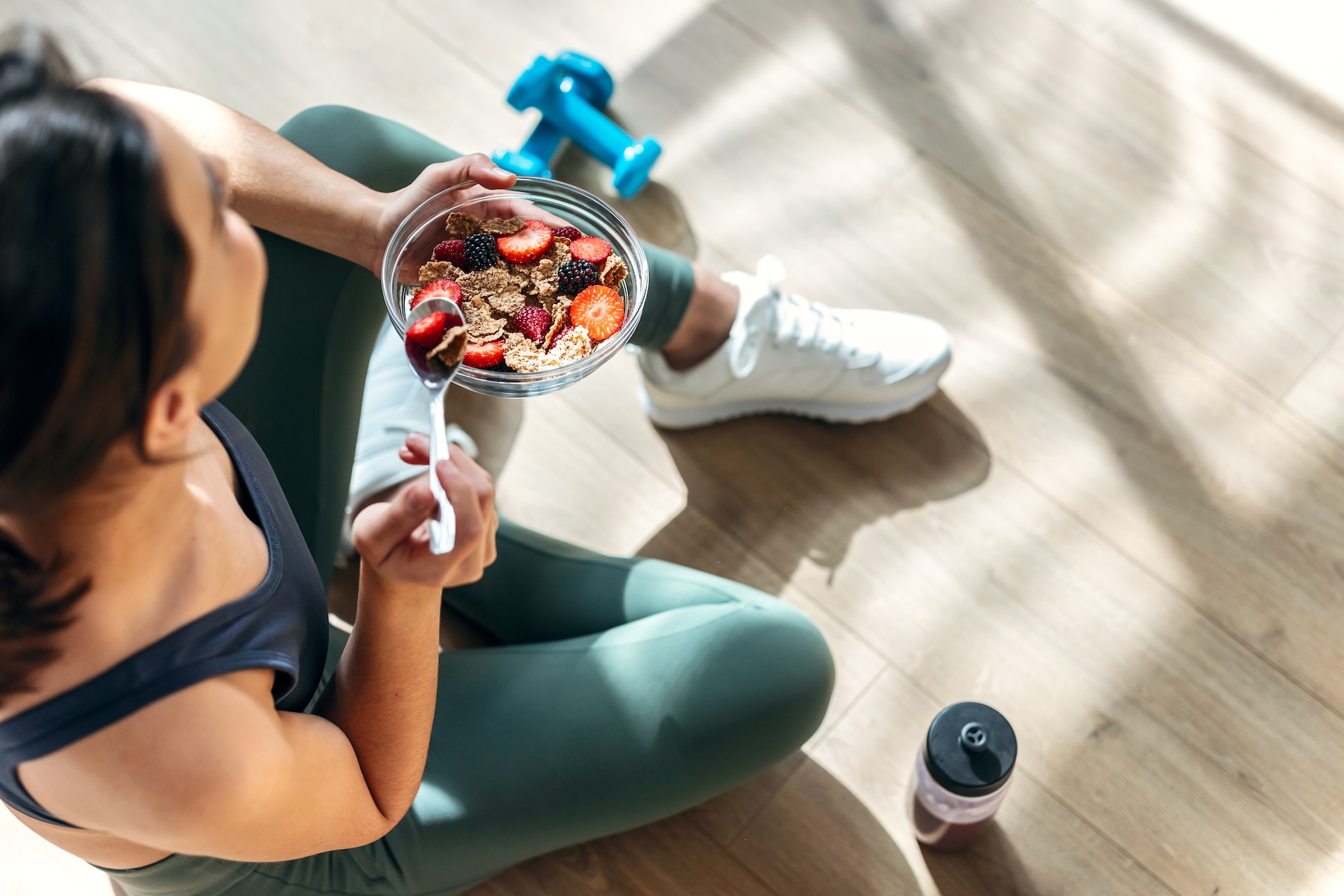 Shot of athletic woman eating a healthy bowl of muesli with fruit sitting on floor in the kitchen at home